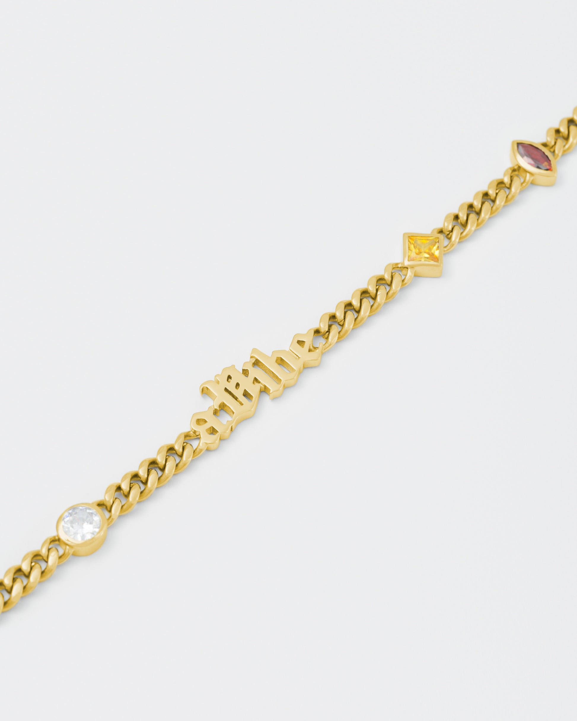 detail of A Vibe cuban chain bracelet with 18kt yellow gold coating, mixed shape bezel stones in diamond white, yellow, garnet, tanzanite and "A Vibe" central metal tag. Lobster clasp with logo