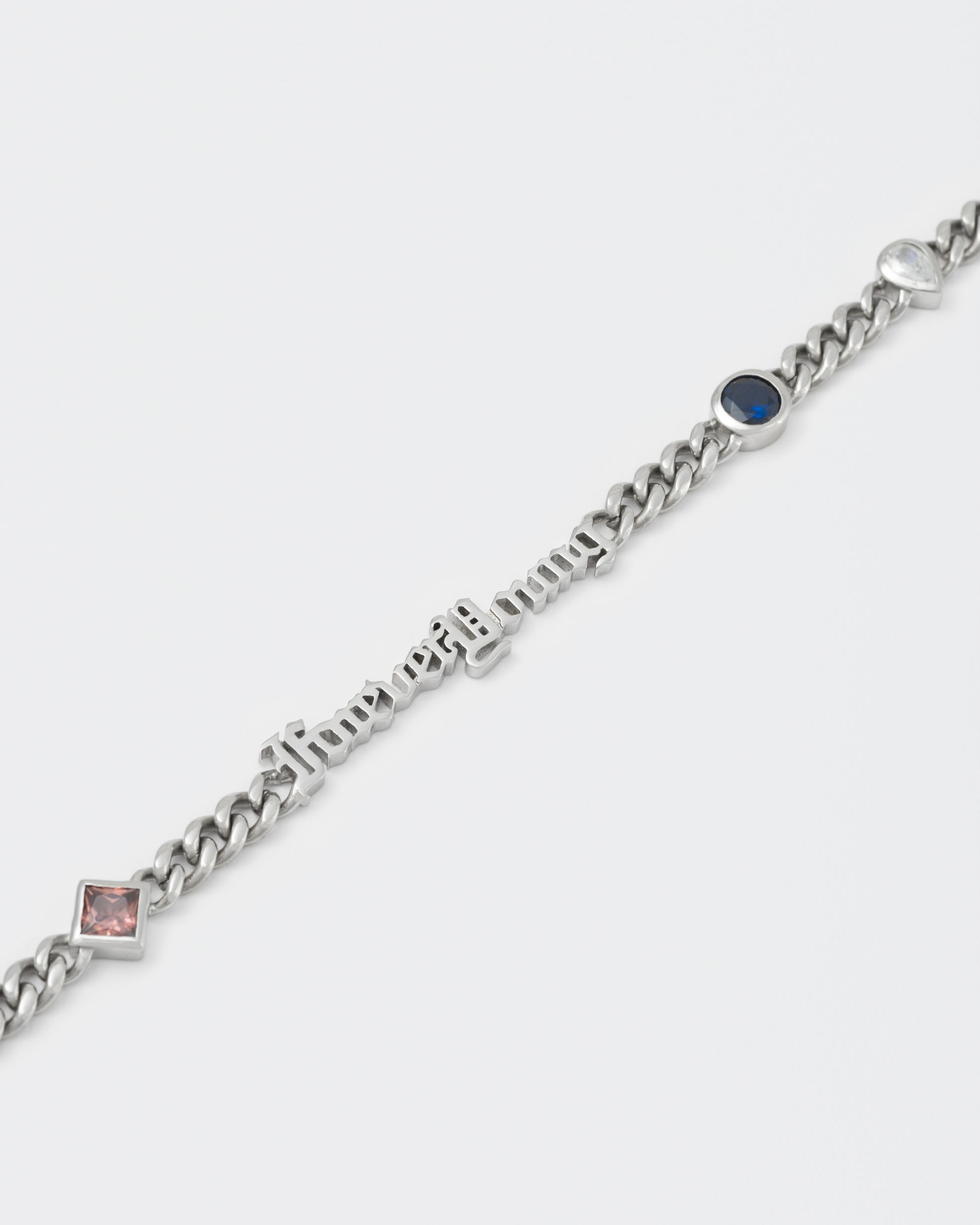 detail of forever Young cuban chain bracelet with 18kt white gold coating, mixed shape bezel stones in diamond white, burna blue, green, chocolate and "Forever Young" central metal tag