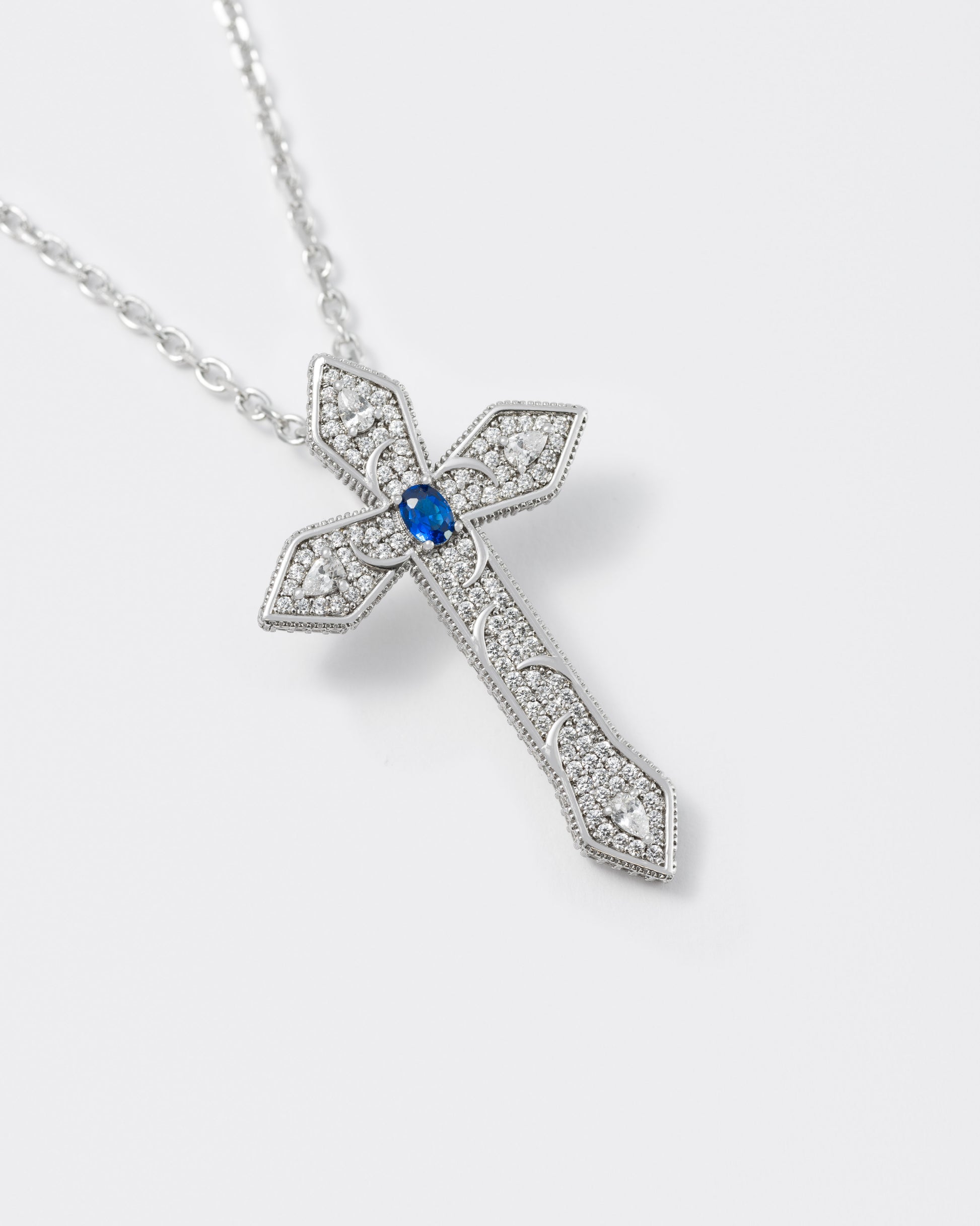 detail of gothic cross pendant necklace with 18kt white gold coating, drop and oval shaped stones in diamond white and sapphire blue with metal gothic detailing. Hand-set micropavé on front and sides