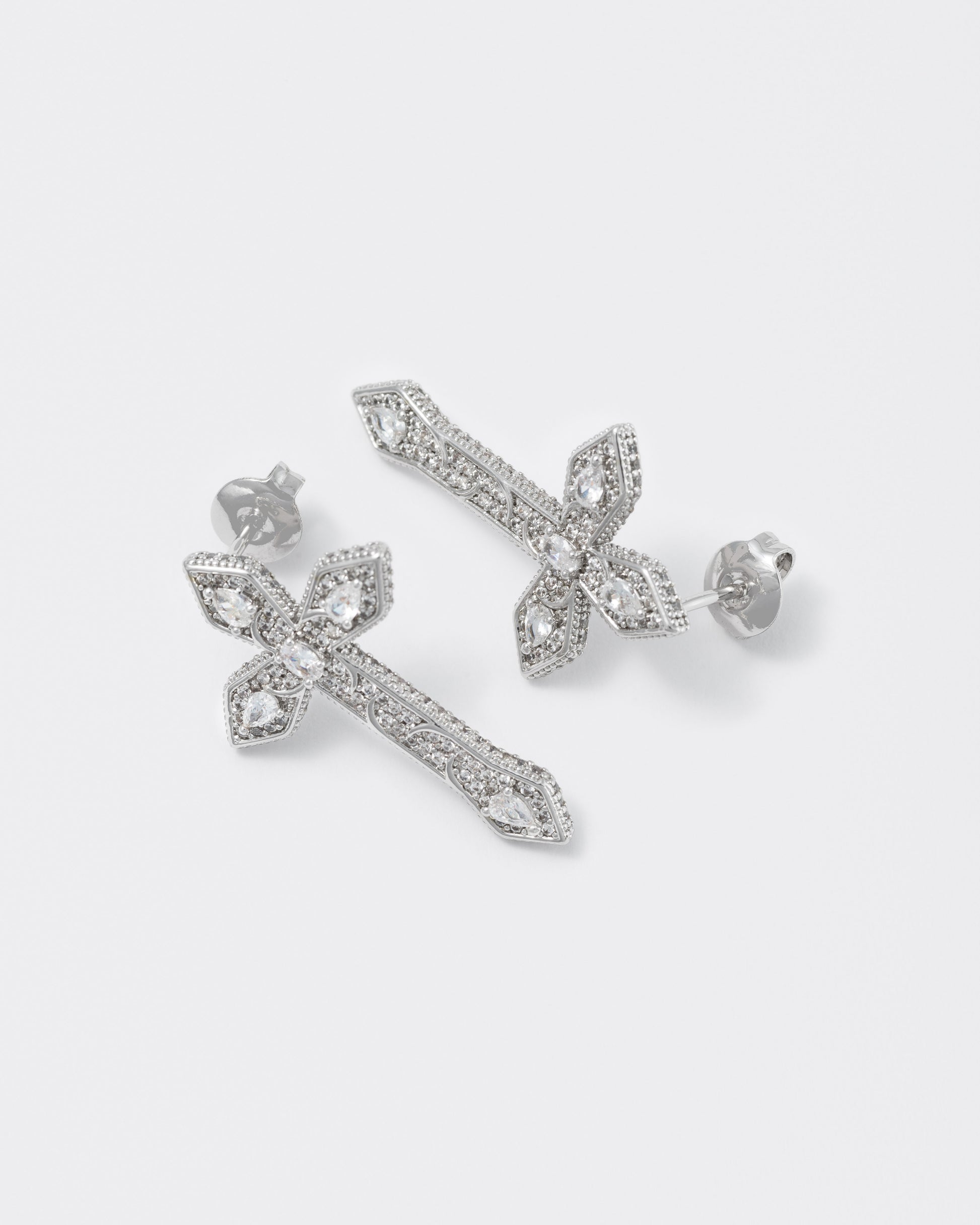 detail of gothic cross earrings with 18kt white gold coating, drop and oval shaped diamond white stones with metal gothic detailing. Hand-set micropavé on front and sides. Silver 925 pin and butterly closure with logo.